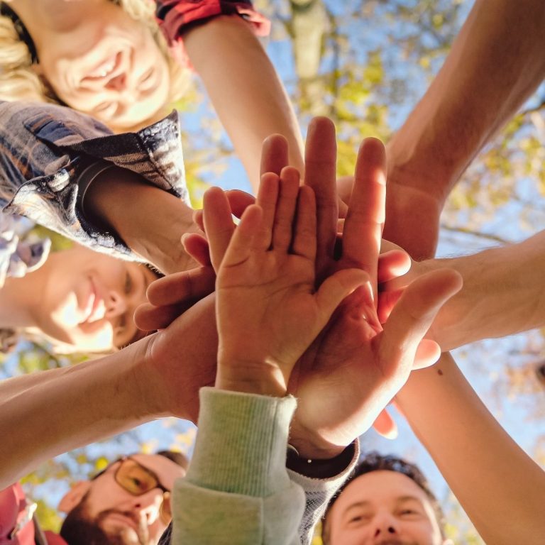 Eine Vielzahl an Händen ist in die Mitte gestreckt, darüber sind glückliche Gesichter zu sehen